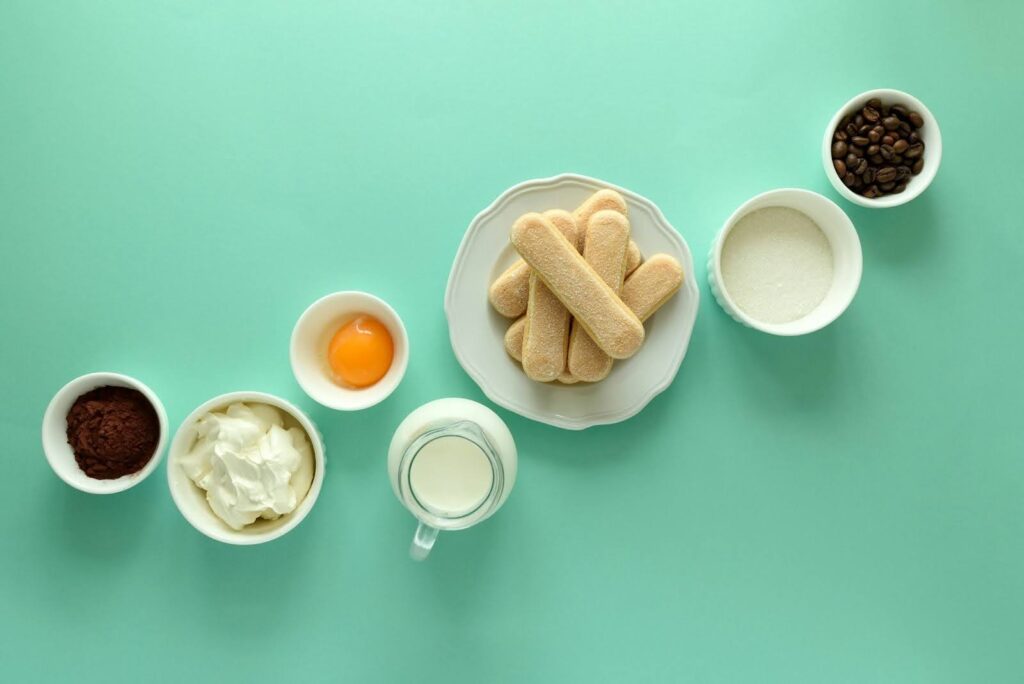 Tiramisu ingredients: ladyfingers, cocoa, mascarpone, egg, milk, sugar, coffee beans on teal background.