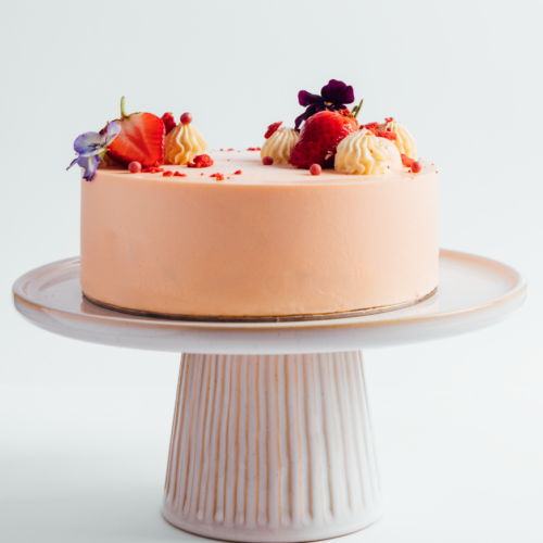 Peach-colored cake with strawberries and flowers on a white stand.