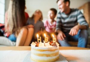 Birthday cake with "HBD" candles, people smiling in the background.