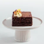 A square chocolate cake with a textured coating, topped with cream, fruit, and edible flowers, displayed on a white cake stand.