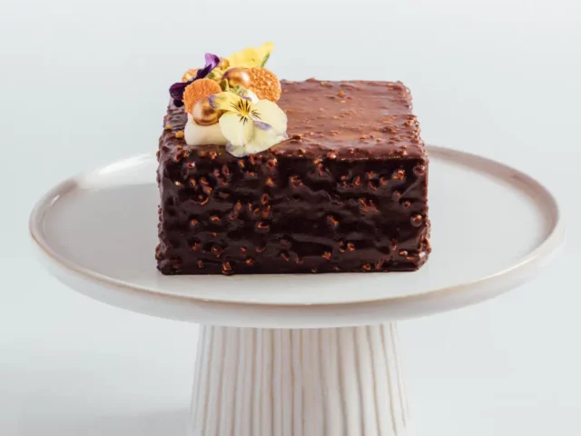 A square chocolate cake with a textured coating, topped with cream, fruit, and edible flowers, displayed on a white cake stand.